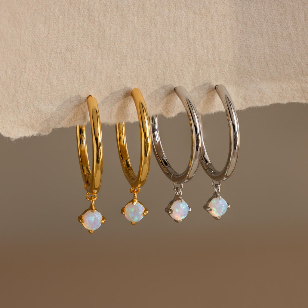 Two pairs of Dainty Opal Drop Huggies—one in gold and one in silver—feature dainty opal drops and rest elegantly on a beige surface.