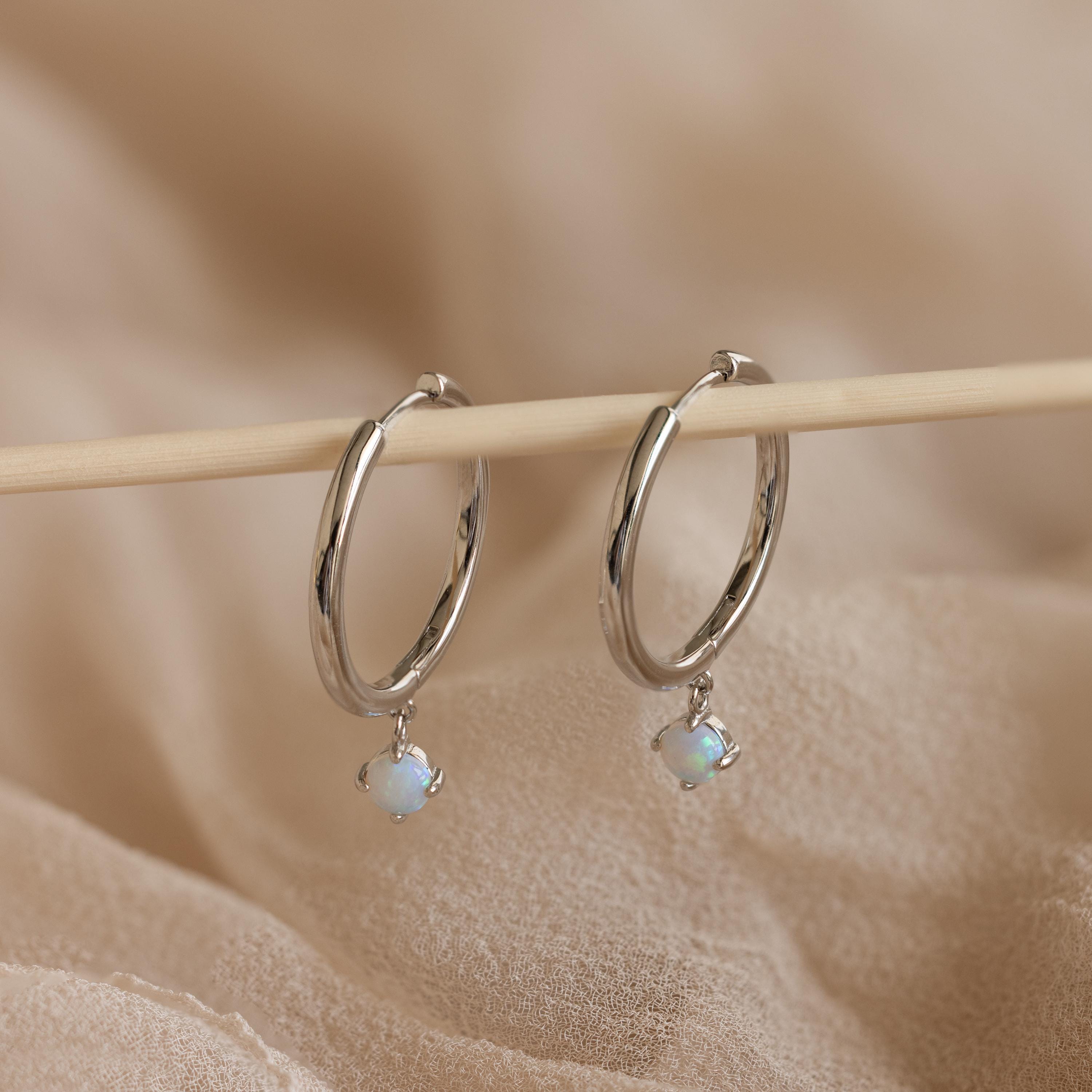 Dainty Opal Drop Huggies feature delicate opal stones on silver hoops, displayed on a wooden stick over beige fabric. These dainty earrings add a subtle shimmer and elegance to any look.