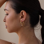 Woman with dark hair in a ponytail wearing Dainty Opal Drop Huggies—delicate gold hoops featuring a petite dangling opal charm.