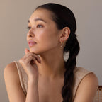 A woman with a long dark braid, wearing Dainty Opal Drop Huggies, gazes to the side with her hand resting thoughtfully on her chin.