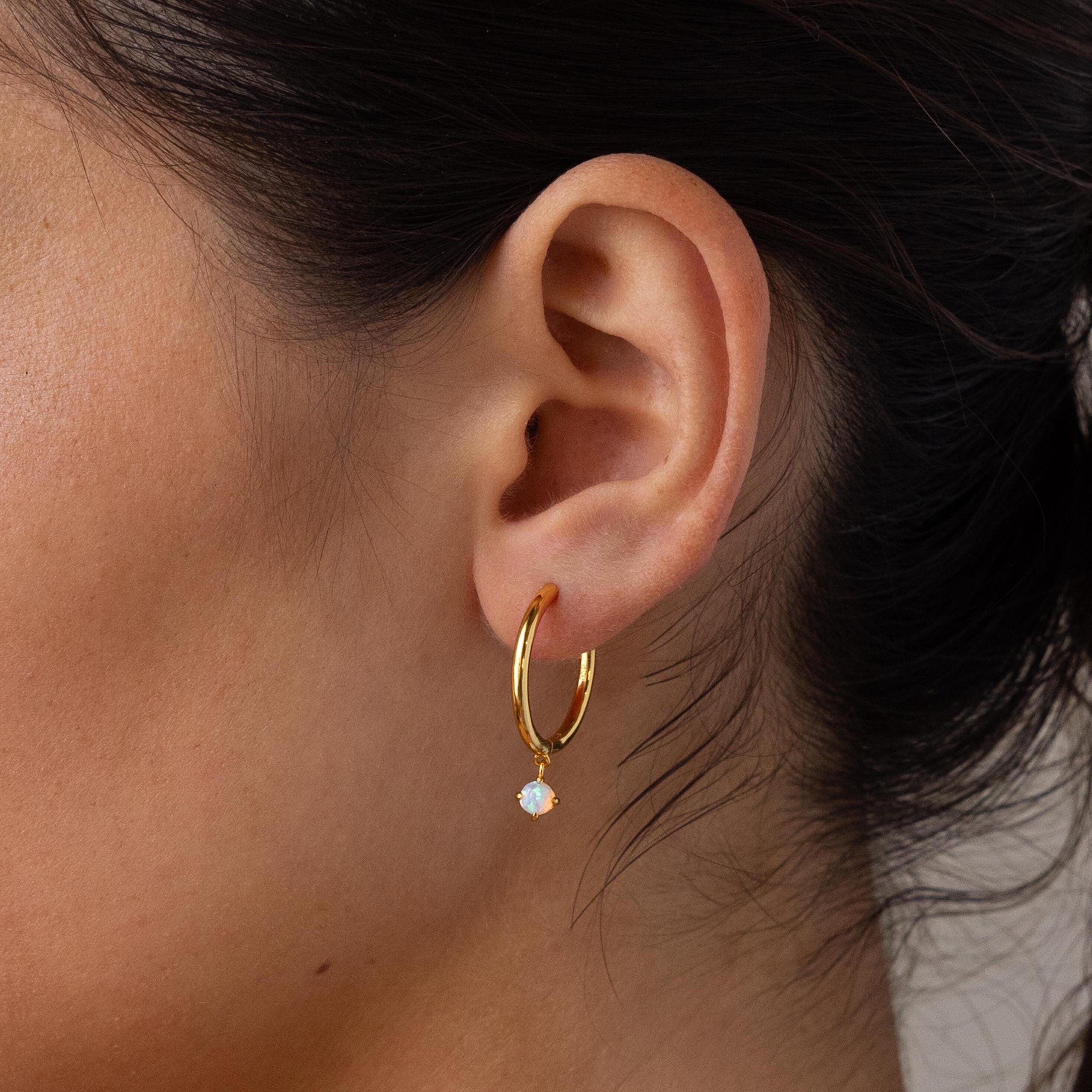 Close-up of a woman's ear wearing Dainty Opal Drop Huggies—gold hoop earrings each with a delicate, dangling opal gemstone.