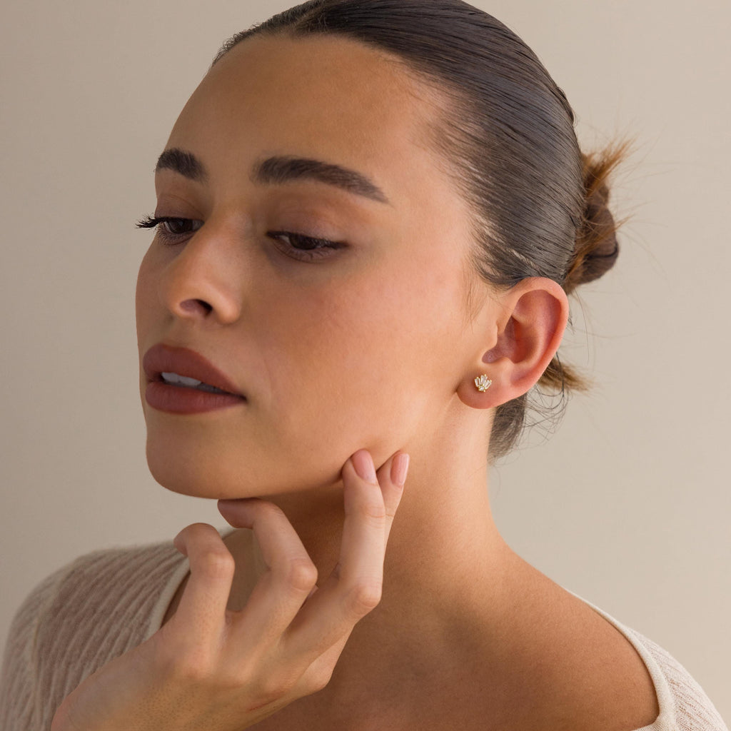 A woman with dark hair in a bun, wearing a beige top, touches her chin while showcasing the Art Deco Studs—gold earrings inspired by vintage glamour.
