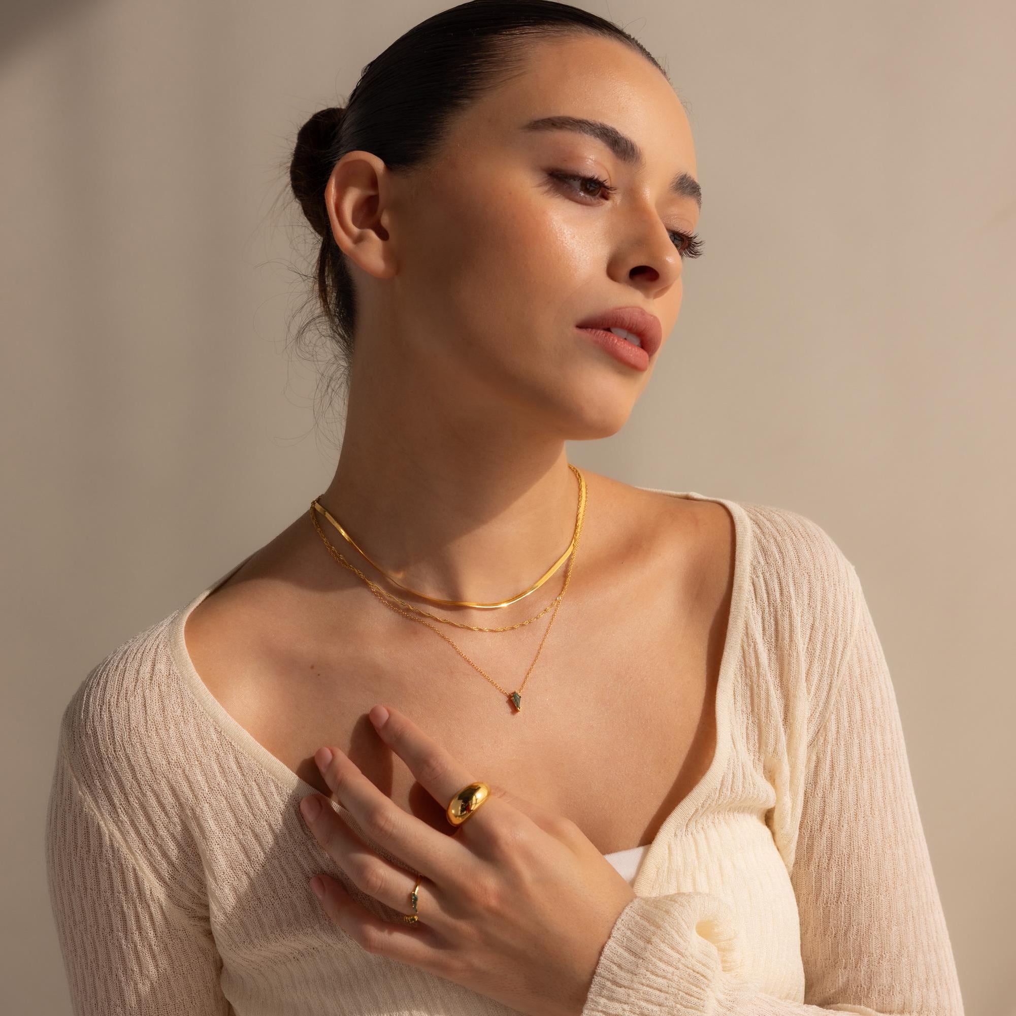 Woman in a cream top layers gold necklaces, including the Agate Art Deco Pendant Necklace, with rings and poses with hand on chest against a neutral background.