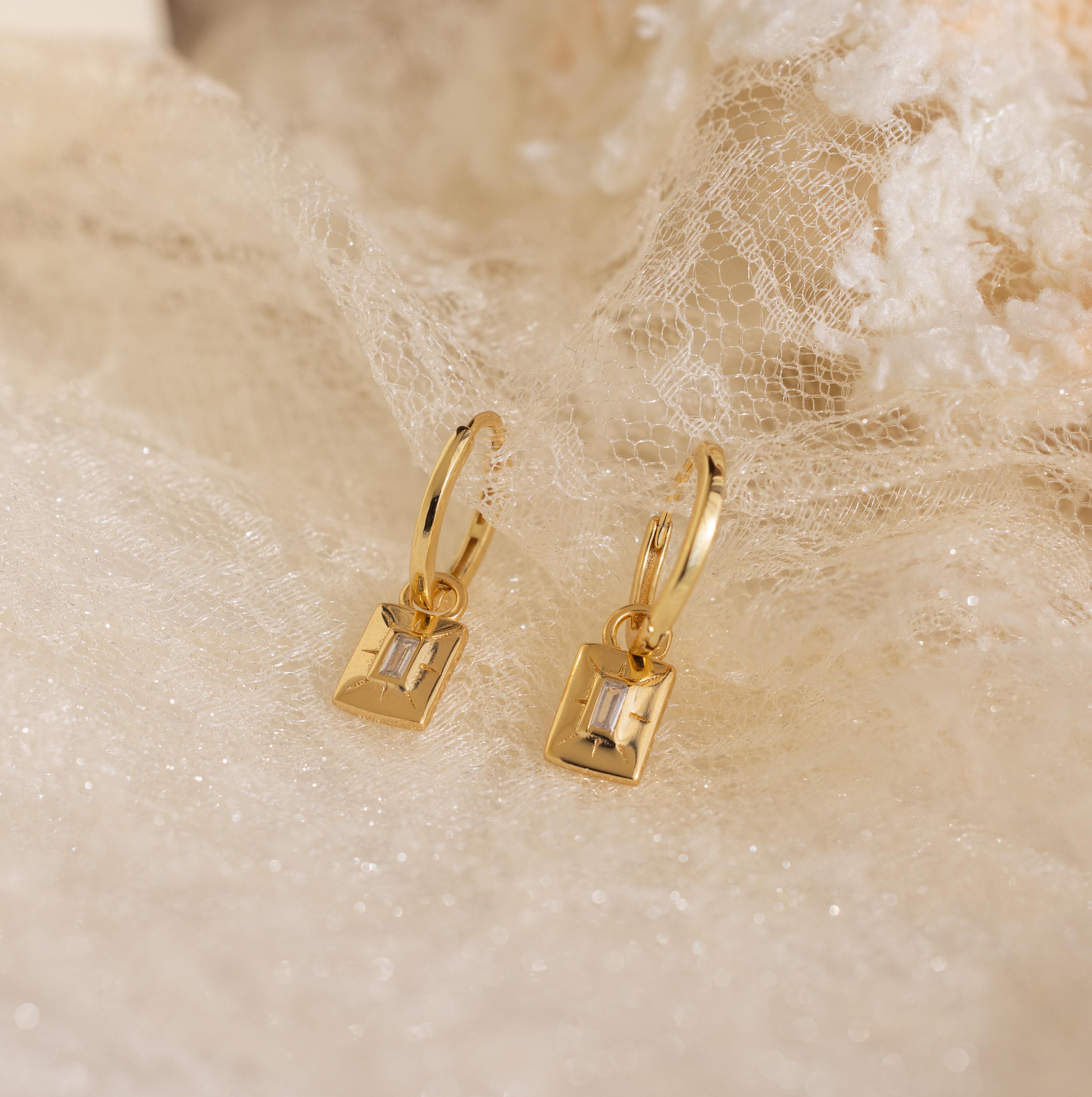 The Art Deco Diamond Tag Huggies showcase rectangular, clear gemstones in vintage-inspired gold hoops, elegantly displayed on delicate lace fabric.