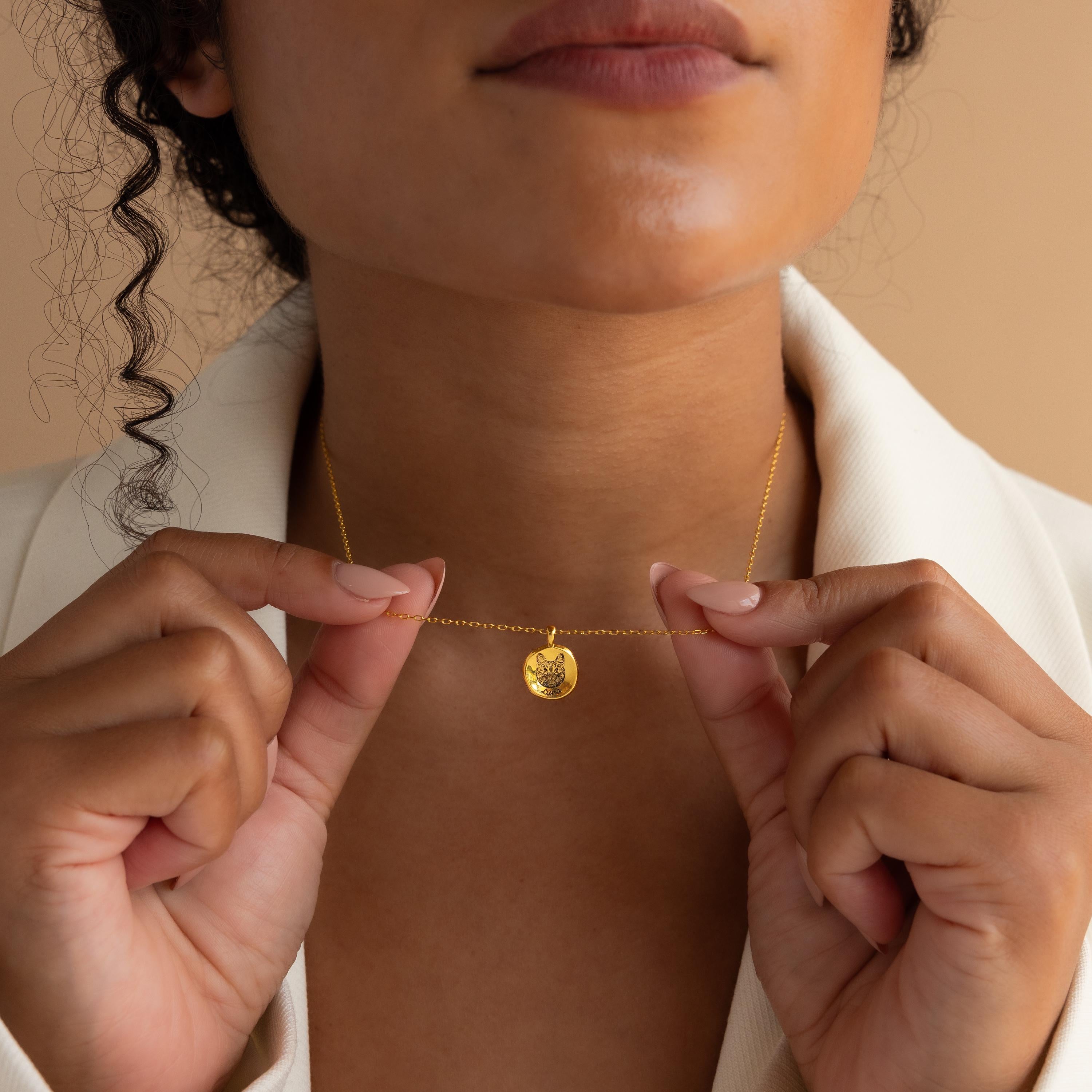 A woman holds the Pet Portrait Coin Necklace—a gold necklace featuring a round, engraved pendant—close to her neck, keeping her beloved companion near in an elegant and personal way.