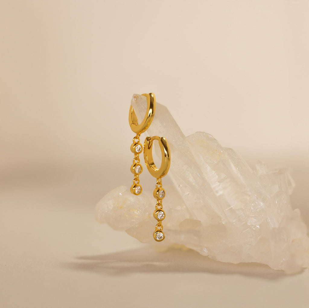 Dangling Diamond Chain Huggies, inspired by art deco jewelry, displayed on a cluster of clear quartz crystals against a beige background.