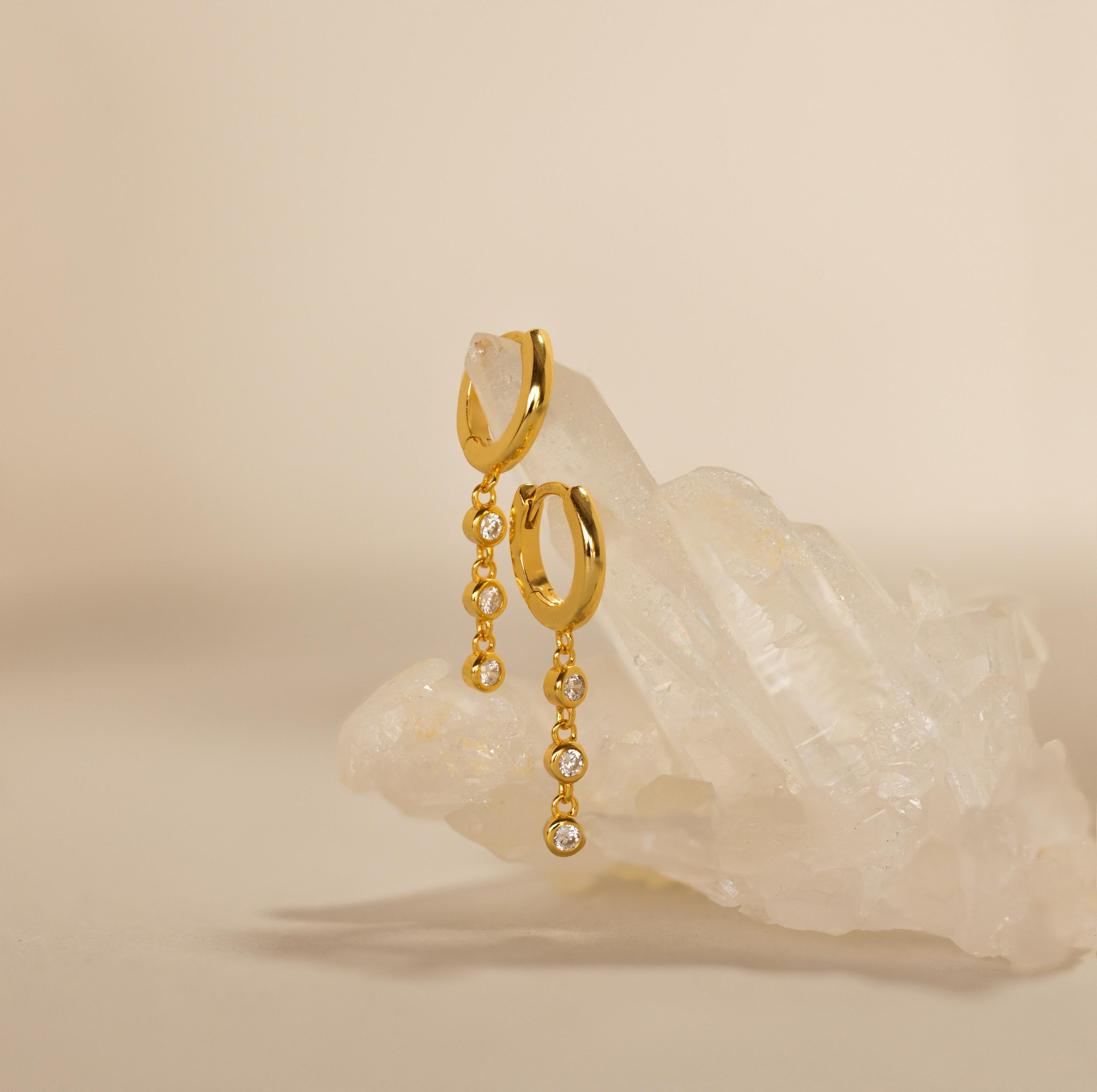 Dangling Diamond Chain Huggies, inspired by art deco jewelry, displayed on a cluster of clear quartz crystals against a beige background.