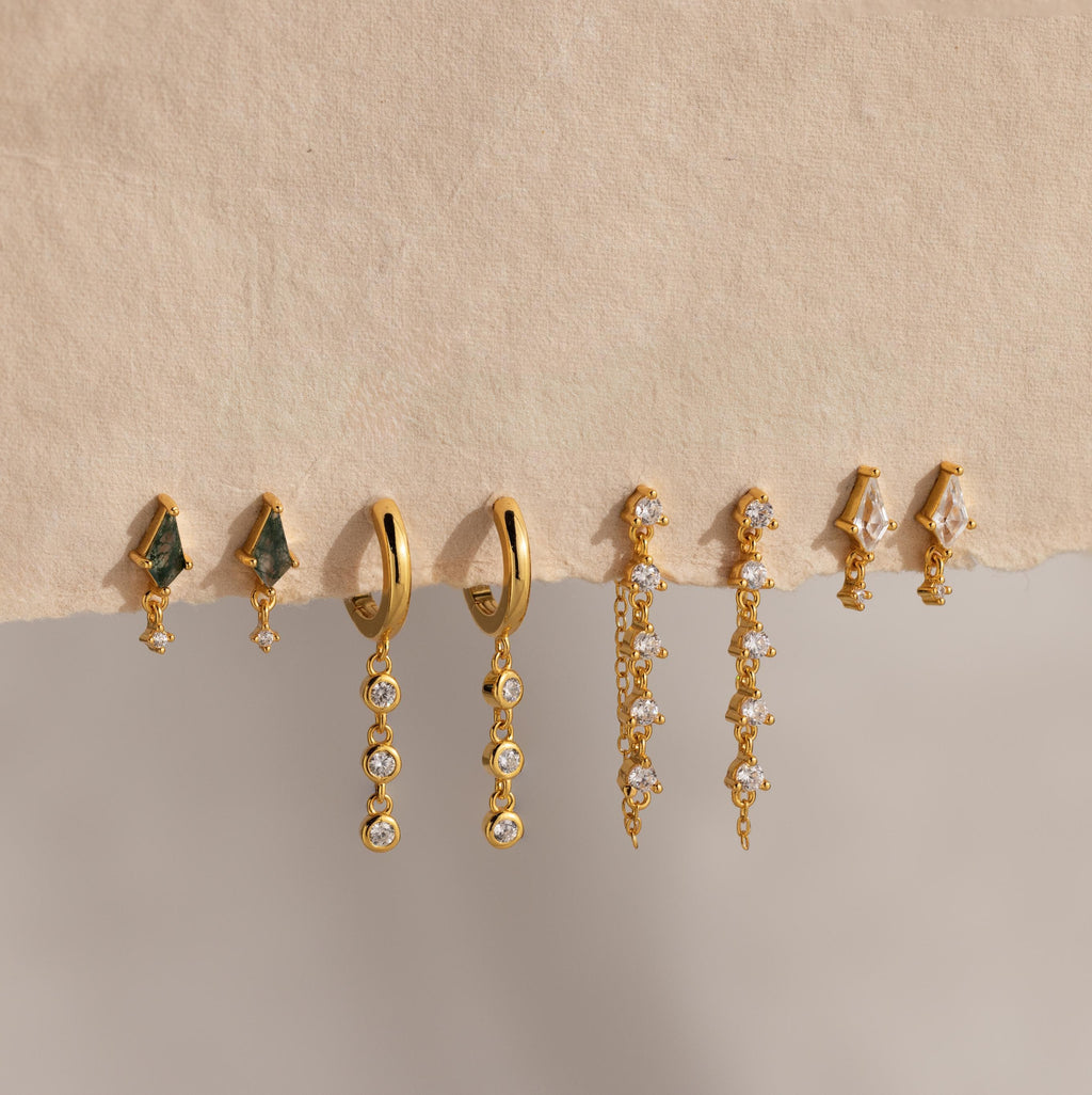 Four pairs of gold and gemstone earrings, featuring Dangling Diamond Chain Huggies, are displayed on a textured beige surface.