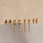 Four pairs of gold and gemstone earrings, featuring Dangling Diamond Chain Huggies, are displayed on a textured beige surface.