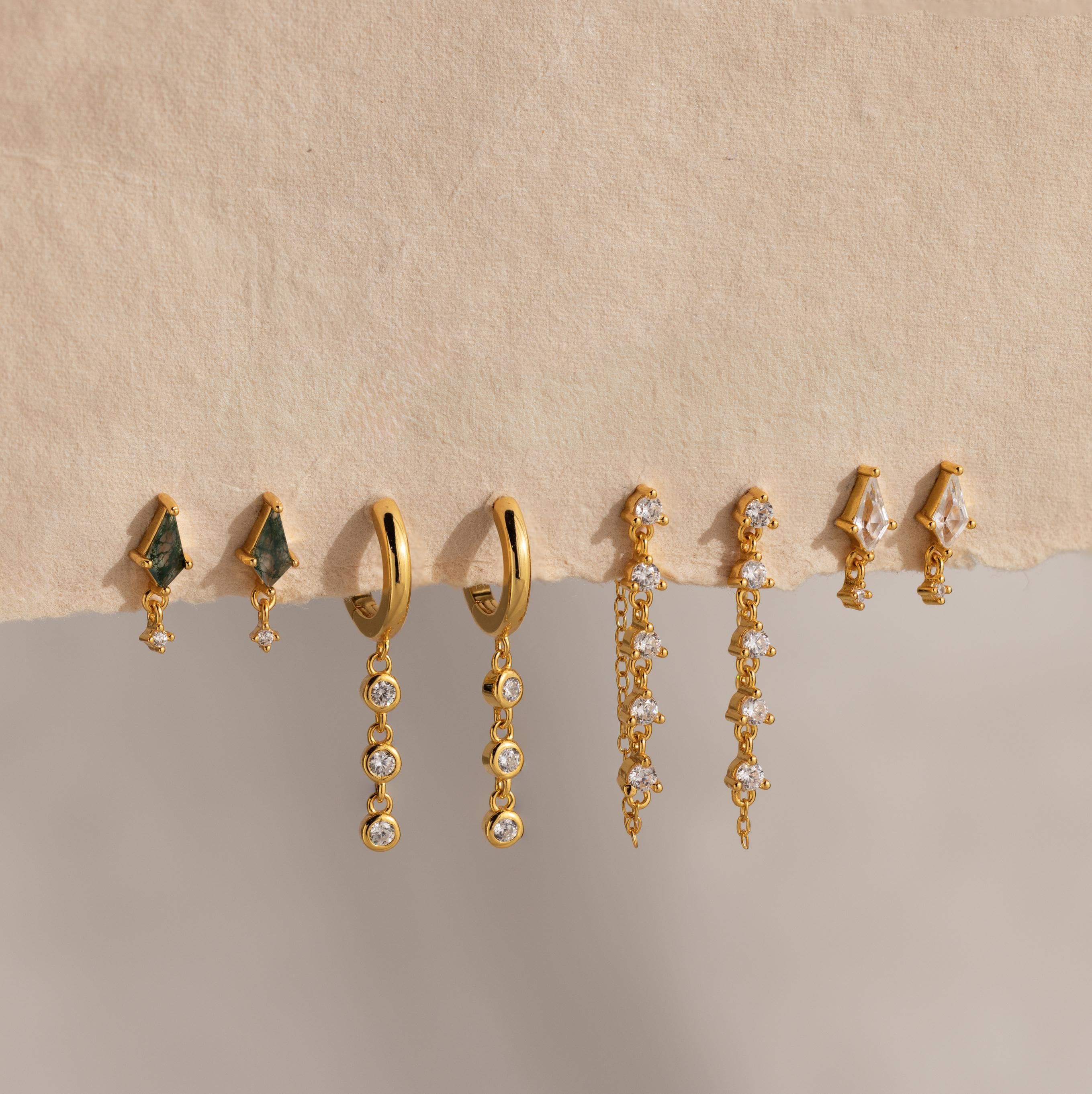 Four pairs of gold and gemstone earrings, featuring Dangling Diamond Chain Huggies, are displayed on a textured beige surface.