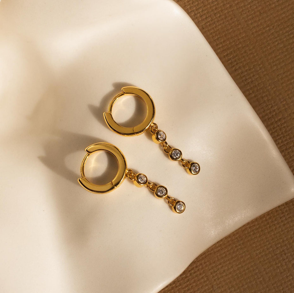 Dangling Diamond Chain Huggies, inspired by art deco style, feature gold hoops with diamond chains and are displayed on cream fabric beside a textured brown surface.