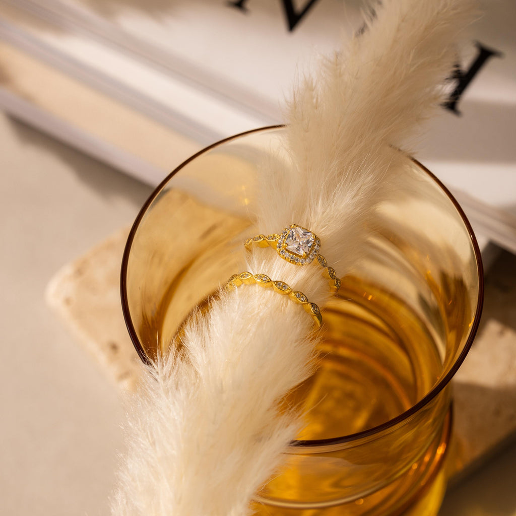 The Vintage Diamond Rings Set, featuring a cushion-cut halo and marquise-detailed band, rests on a fluffy white stem inside an amber glass cup with blurred books in the background.