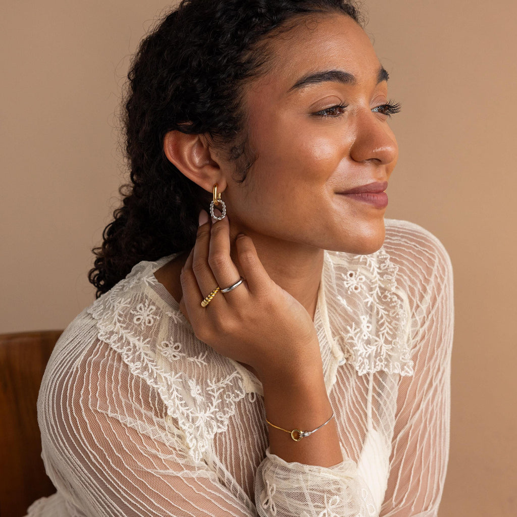 Woman in a lace top smiles, touching her Mixed Metal Link Hoops and wearing rings and a bracelet against a beige background.