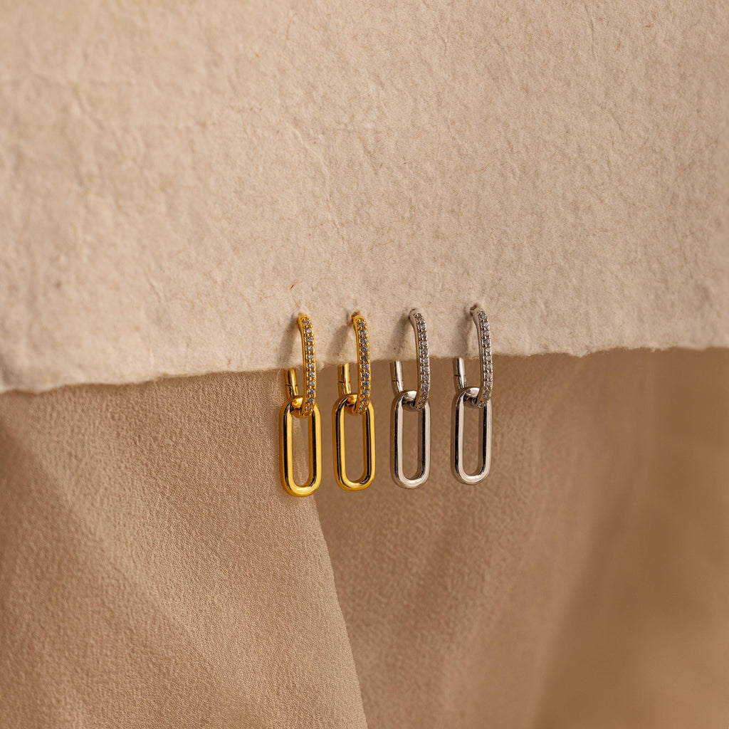 Two pairs of Pave Link Drop Earrings, one in gold and one in silver, rest on a textured beige surface. Each pair sparkles with elegant CZ Diamonds, capturing the classic beauty of rectangular hoop pave link drop earrings.