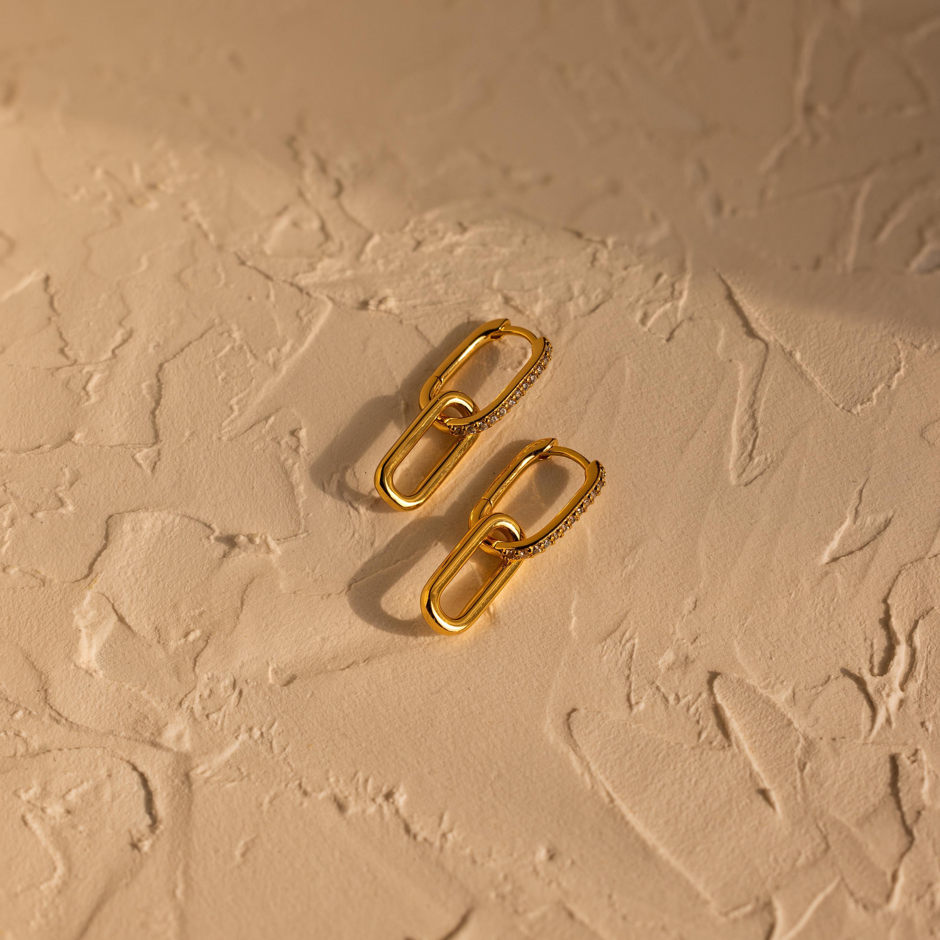 Pave Link Drop Earrings feature gold geometric links and sparkling CZ diamonds, displayed on a textured beige surface with soft shadows.