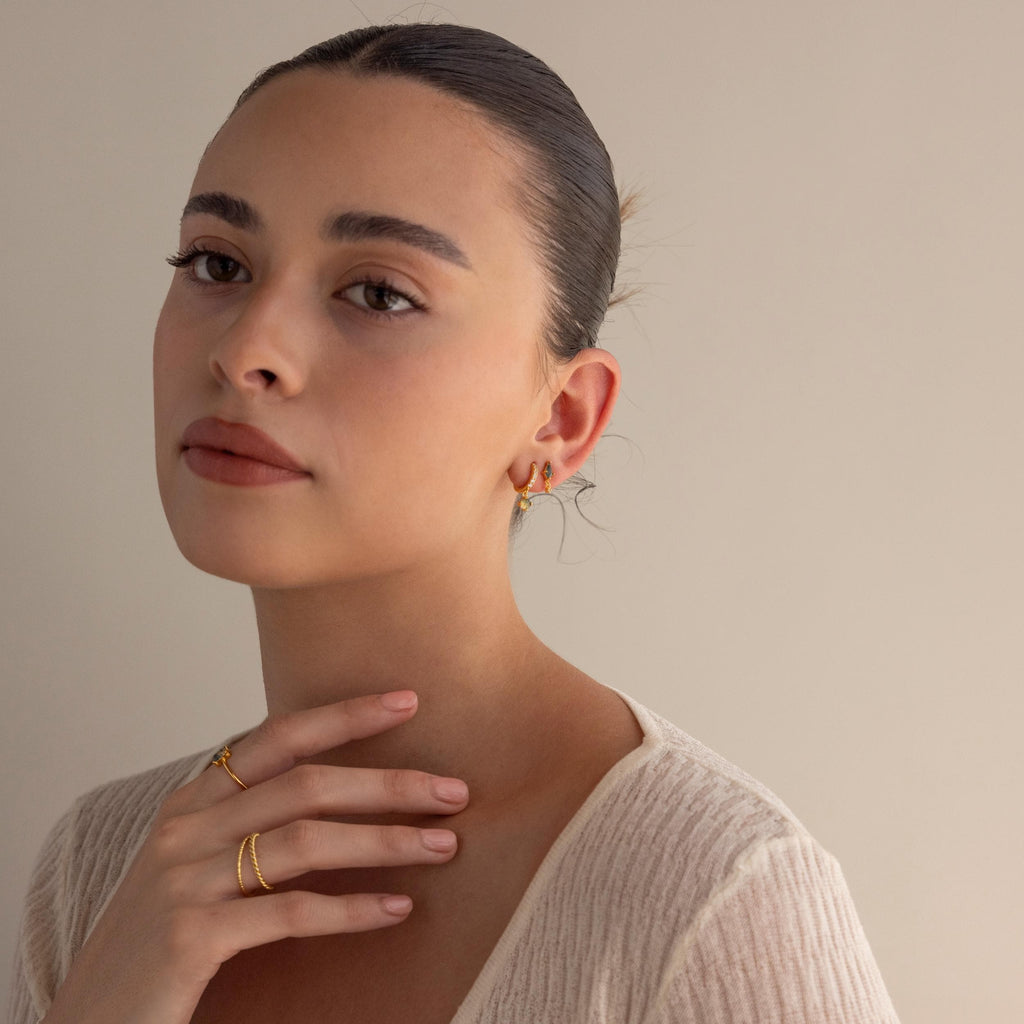 A woman with slicked-back hair, wearing gold rings and Agate Kite Drop Earrings, touches her neck while dressed in a cream top against a beige background.