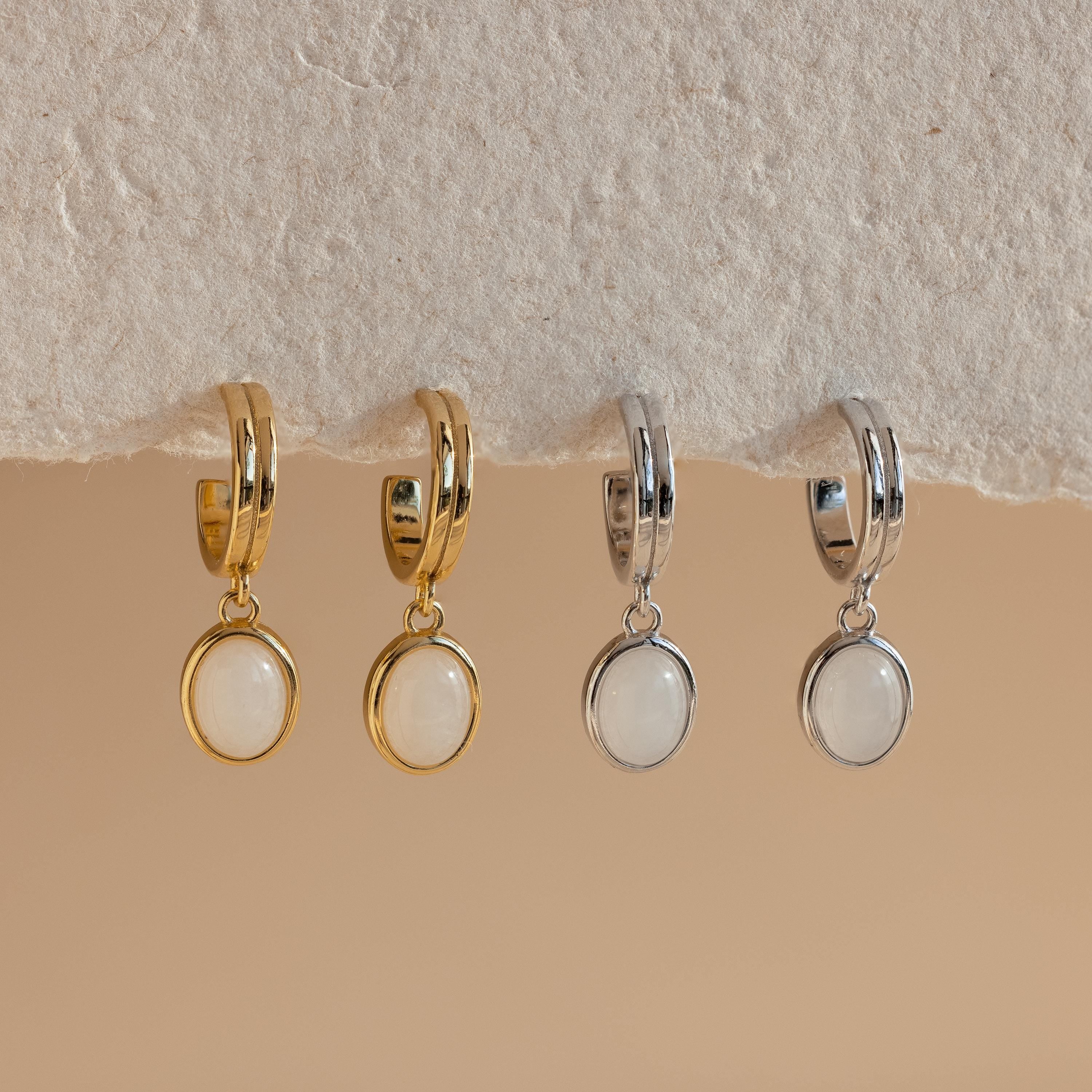 The Moonstone Drop Hoops, available in gold and silver with round white stone pendants, evoke celestial vibes—perfect as moonstone earrings for any occasion. Shown displayed on a textured surface.