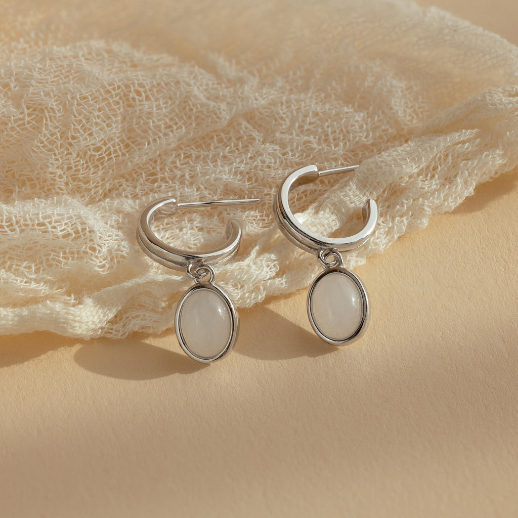 Moonstone Drop Hoops feature silver hoops with oval white pendants, capturing celestial elegance. Displayed on textured cream fabric, these earrings add a touch of moonstone-inspired charm to any look.