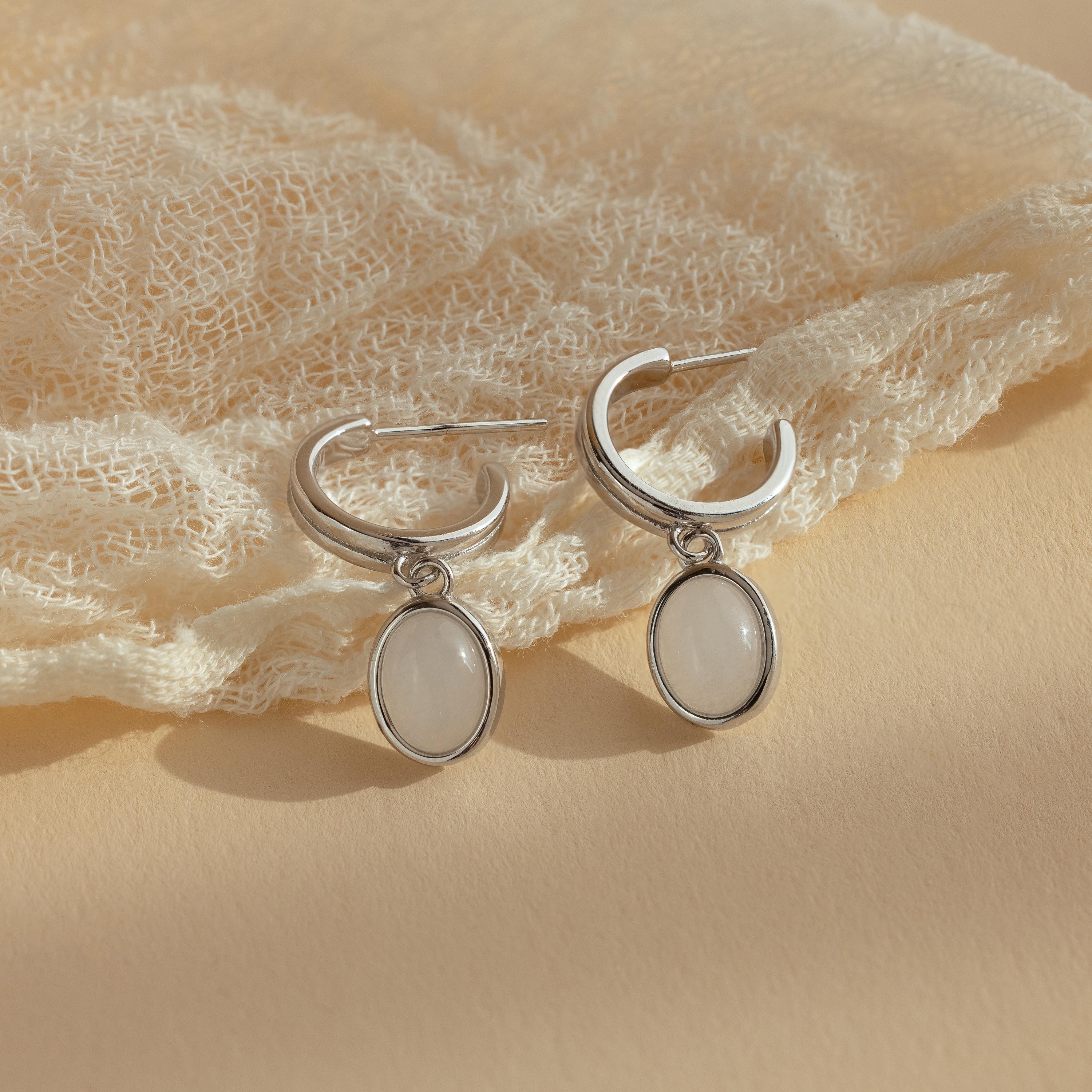 Moonstone Drop Hoops feature silver hoops with oval white pendants, capturing celestial elegance. Displayed on textured cream fabric, these earrings add a touch of moonstone-inspired charm to any look.
