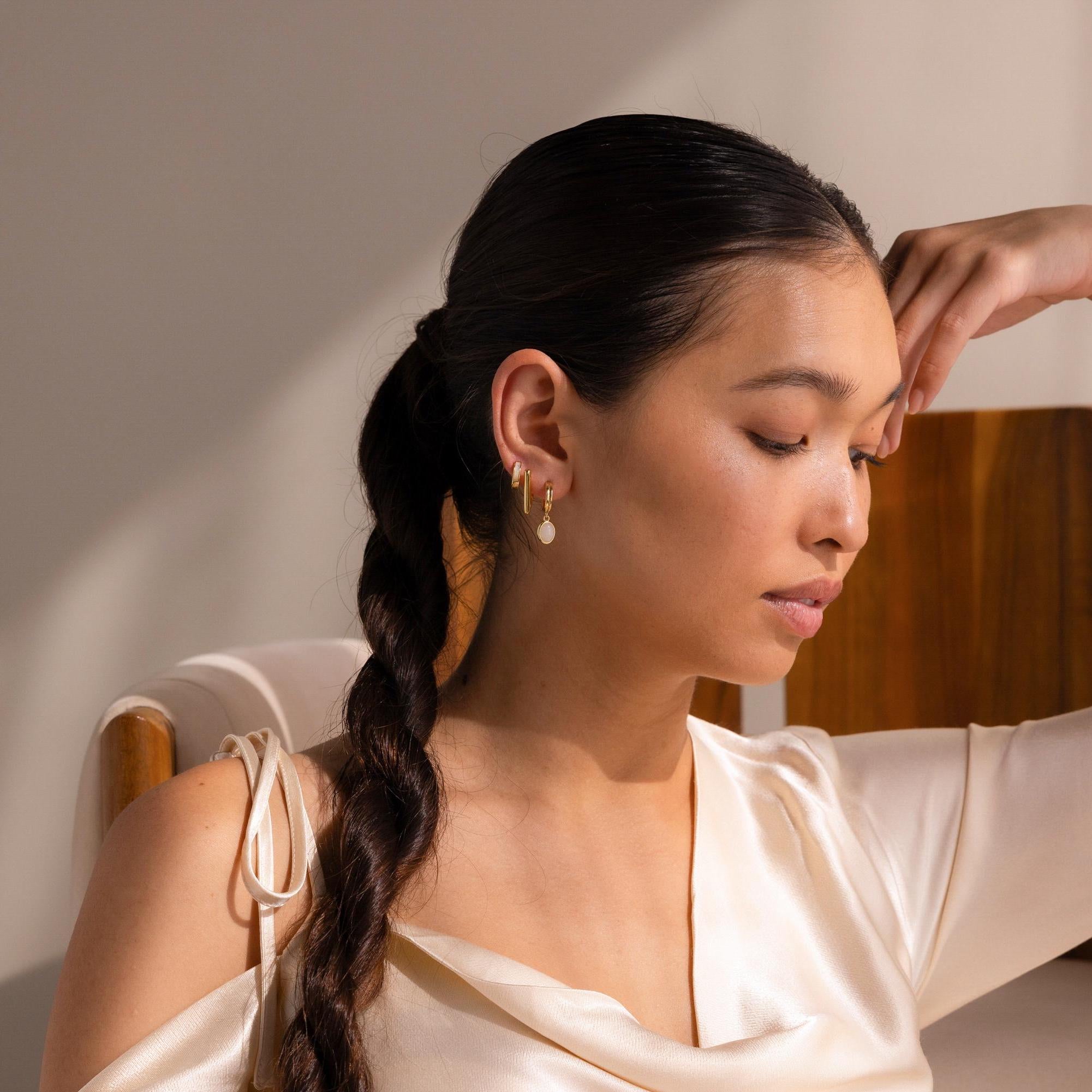 A woman with a long dark braid and gold earrings sits serenely in a chair, wearing a cream satin top. Her eyes are closed as her Moonstone Drop Hoops cast a gentle, celestial glow, making her jewelry softly shimmer.