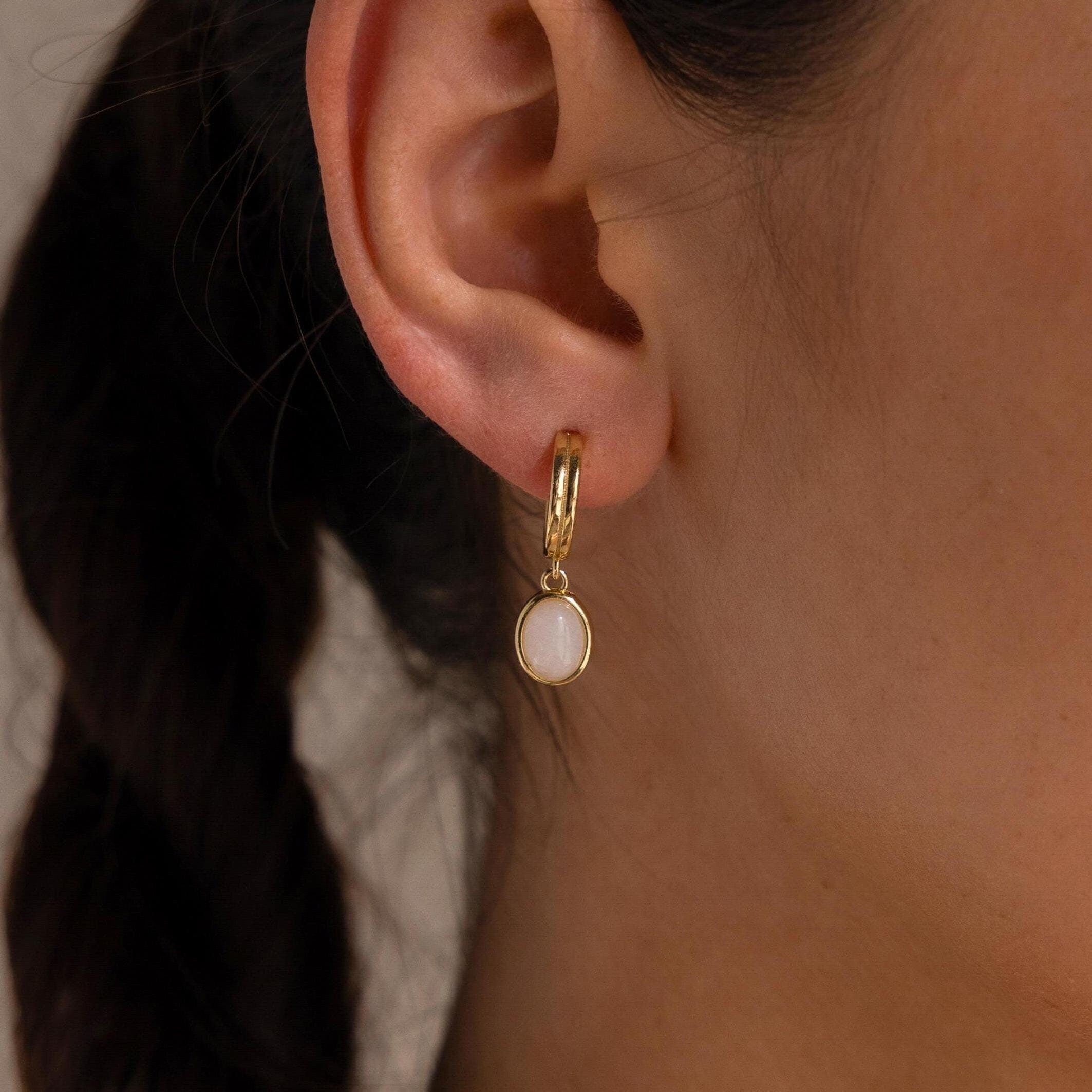 Close-up of an ear wearing a small gold Moonstone Drop Hoops earring featuring a white oval stone pendant, ideal for those who love celestial jewelry.