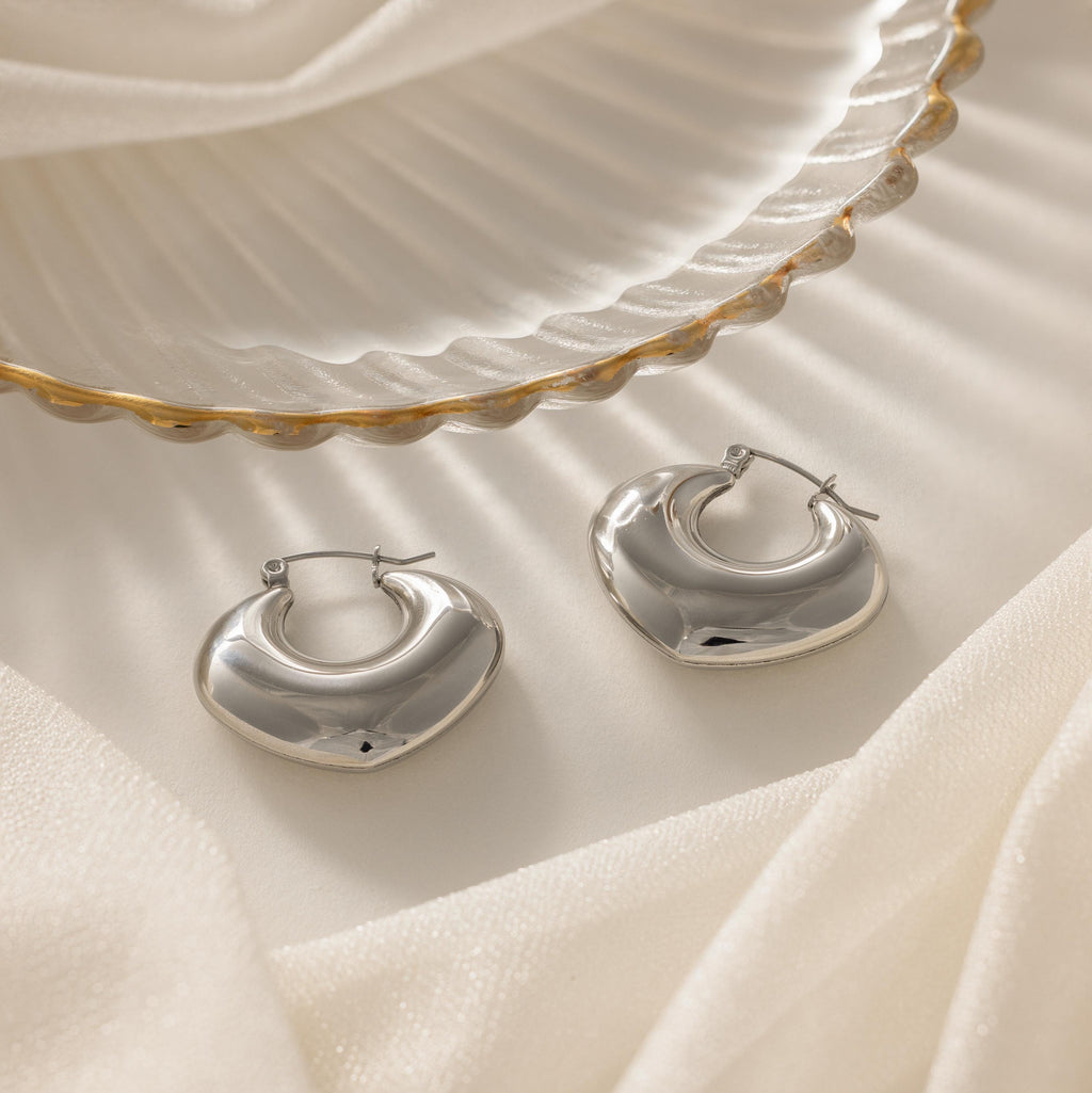 The Chunky Curvy Hoops are shown on a white fabric background beside a scalloped edge dish—ideal minimalist earrings for fans of modern jewelry.