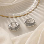 The Chunky Curvy Hoops are shown on a white fabric background beside a scalloped edge dish—ideal minimalist earrings for fans of modern jewelry.
