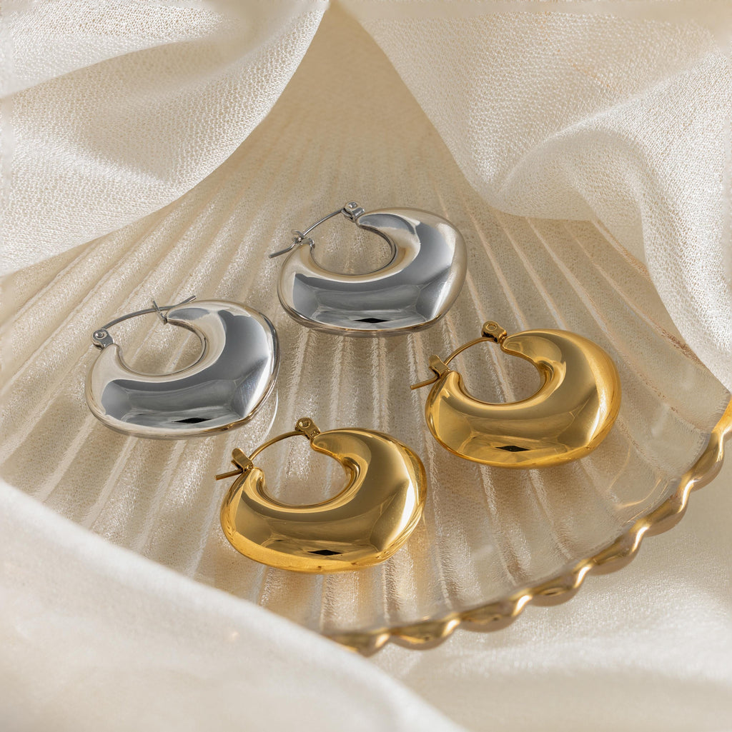 Two pairs of Chunky Curvy Hoops—one gold, one silver—add modern style to your look, elegantly displayed on a shell-shaped dish with fabric for a striking yet minimalist earrings aesthetic.