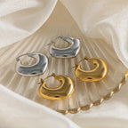 Two pairs of Chunky Curvy Hoops—one gold, one silver—add modern style to your look, elegantly displayed on a shell-shaped dish with fabric for a striking yet minimalist earrings aesthetic.