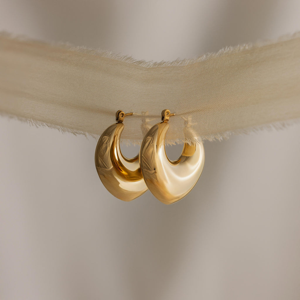 Two shiny gold Chunky Curvy Hoops hang on sheer beige fabric with frayed edges, ideal for fans of minimalist earrings and modern jewelry.