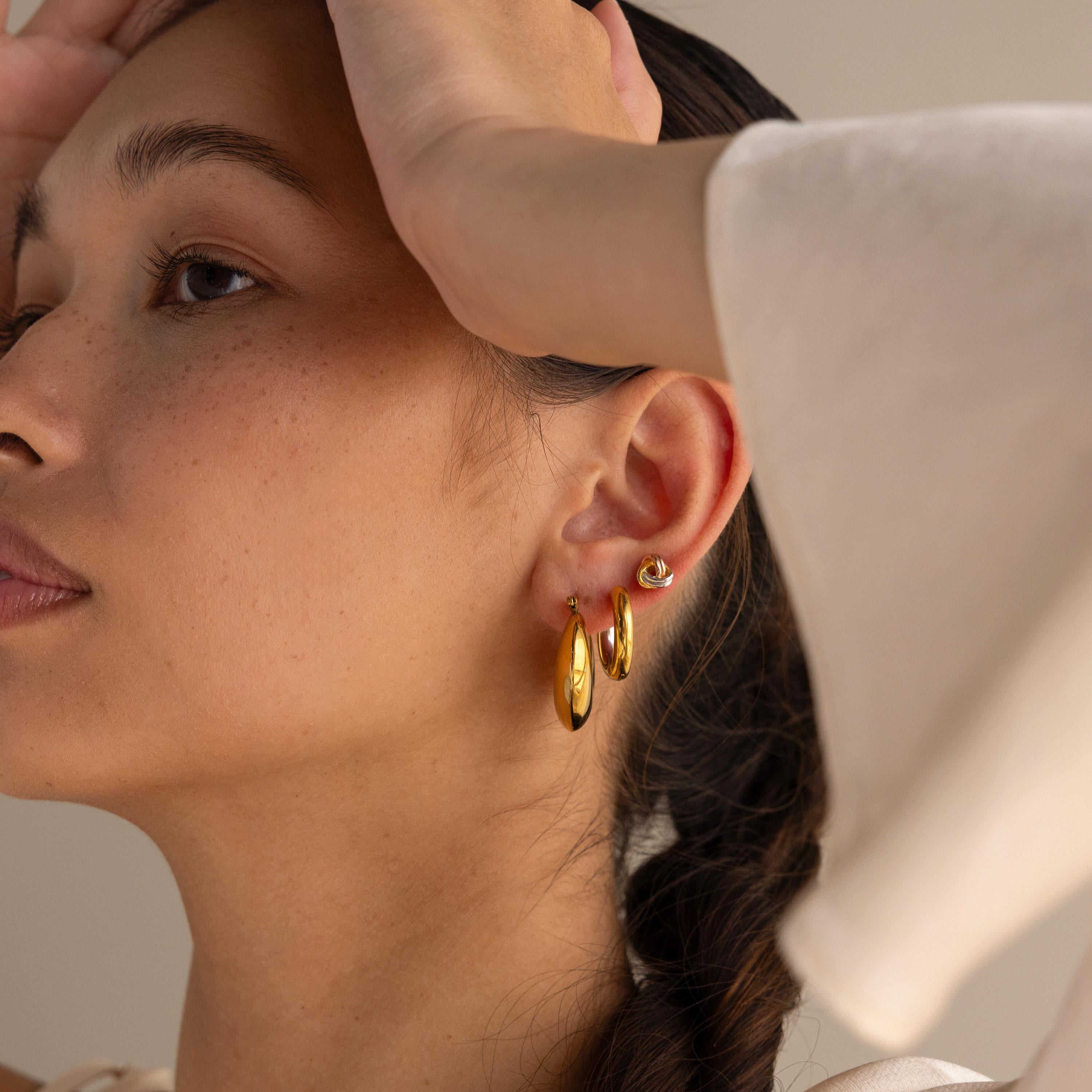 A woman with braided hair wearing Chunky Curvy Hoops lifts her arms to her forehead, bathed in soft light.