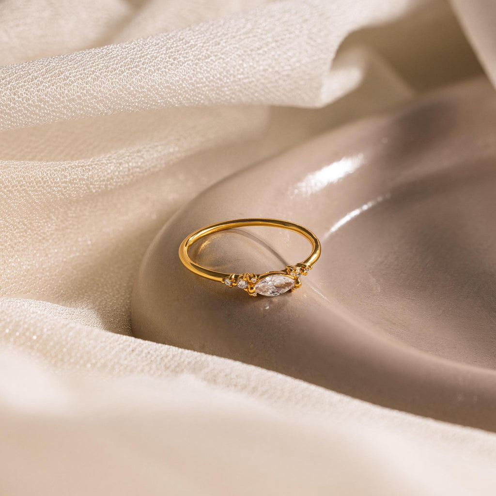 The Marquise Diamond Ring, featuring a gold finish band, is displayed on a smooth beige dish with a soft fabric backdrop.