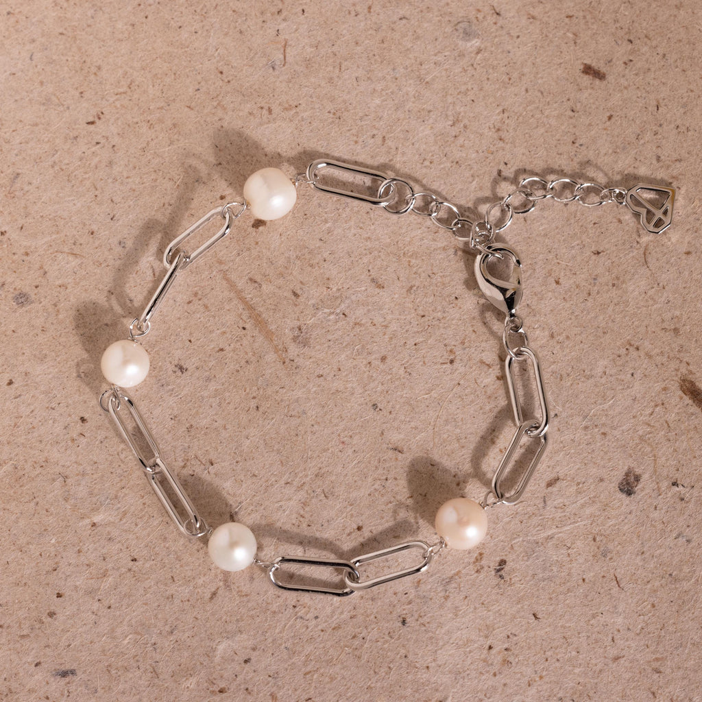 The Pearl Station Paperclip Bracelet features four white pearls on a delicate paperclip chain, beautifully displayed on a textured beige surface—an elegant homage to timeless birthstone jewelry.