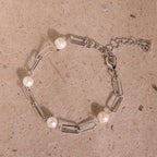 The Pearl Station Paperclip Bracelet features four white pearls on a delicate paperclip chain, beautifully displayed on a textured beige surface—an elegant homage to timeless birthstone jewelry.