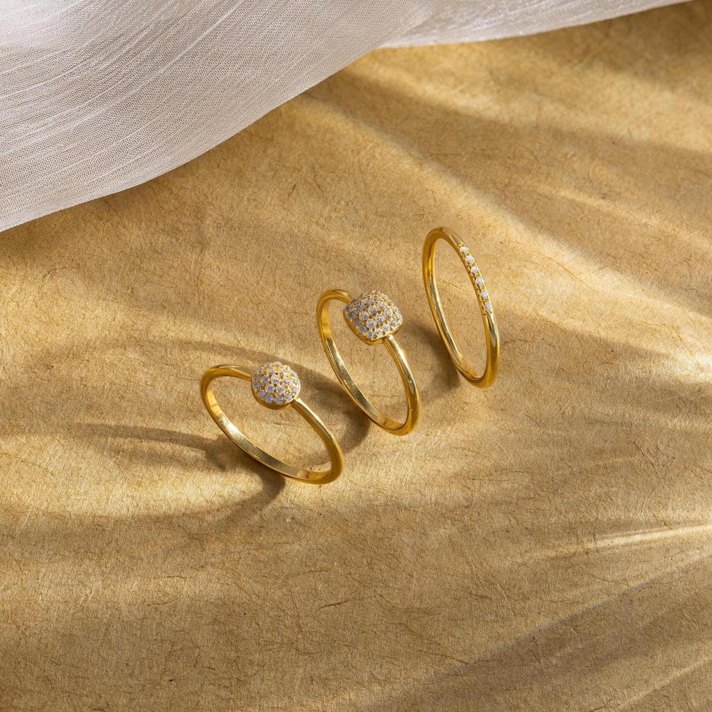 The Classic Pave Ring Set features three gold rings with sparkling stone-studded designs, beautifully displayed on a textured golden surface, adding elegance and sophistication to any jewelry collection.