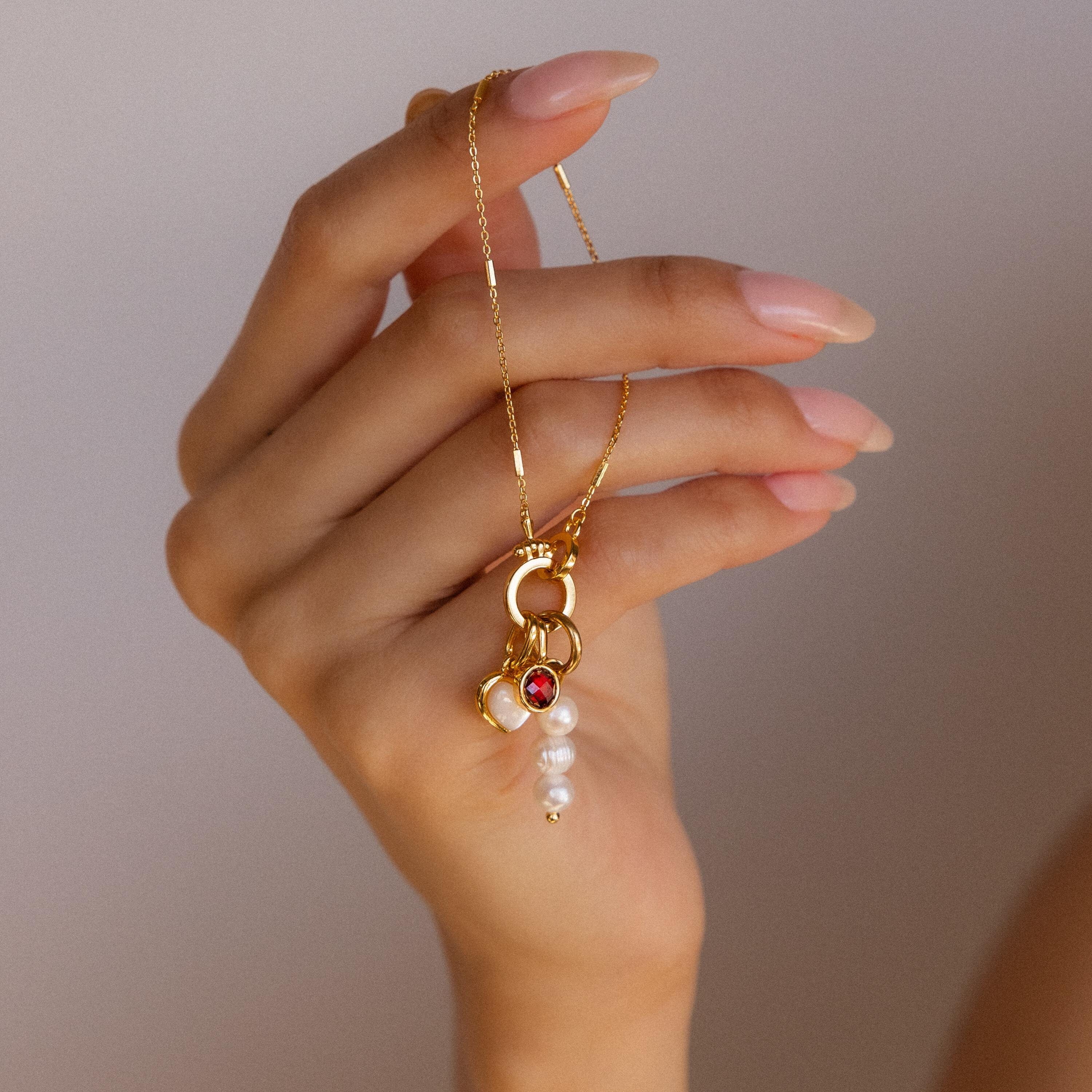 A manicured hand holds the Hinged Circle Charm Bracelet in 18K Gold, featuring delicate red gem and pearl charms that embody the elegance of minimalist jewelry.