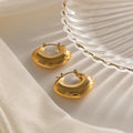 Two shiny Chunky Curvy Hoops in gold rest on soft cream fabric next to a clear, textured glass dish, bringing modern jewelry elegance to the scene.