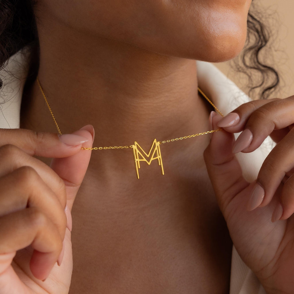 A woman displays the Modern Name Monogram Necklace with an "M" pendant near her neck, highlighting her manicured nails—a chic, personalized jewelry piece.