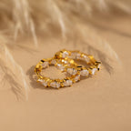 The Inside Out Pear Diamond Hoops, gold statement earrings featuring marquise-cut clear stones, rest elegantly on a beige surface with a blurred plant behind—an ideal bridesmaid gift.