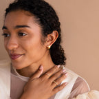 Woman with curly hair wearing Pave Checkered Huggies and rings, touching her neck against a beige background.