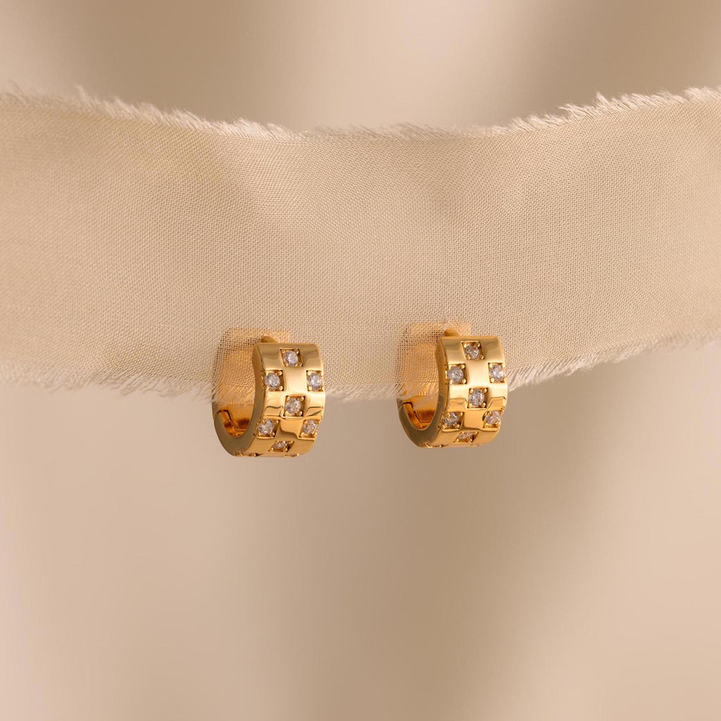 The Pave Checkered Huggies are gold chunky hoops featuring square designs and small gemstones, shown on a sheer beige fabric background.