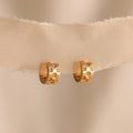 The Pave Checkered Huggies are gold chunky hoops featuring square designs and small gemstones, shown on a sheer beige fabric background.