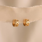 The Pave Checkered Huggies are gold chunky hoops featuring square designs and small gemstones, shown on a sheer beige fabric background.