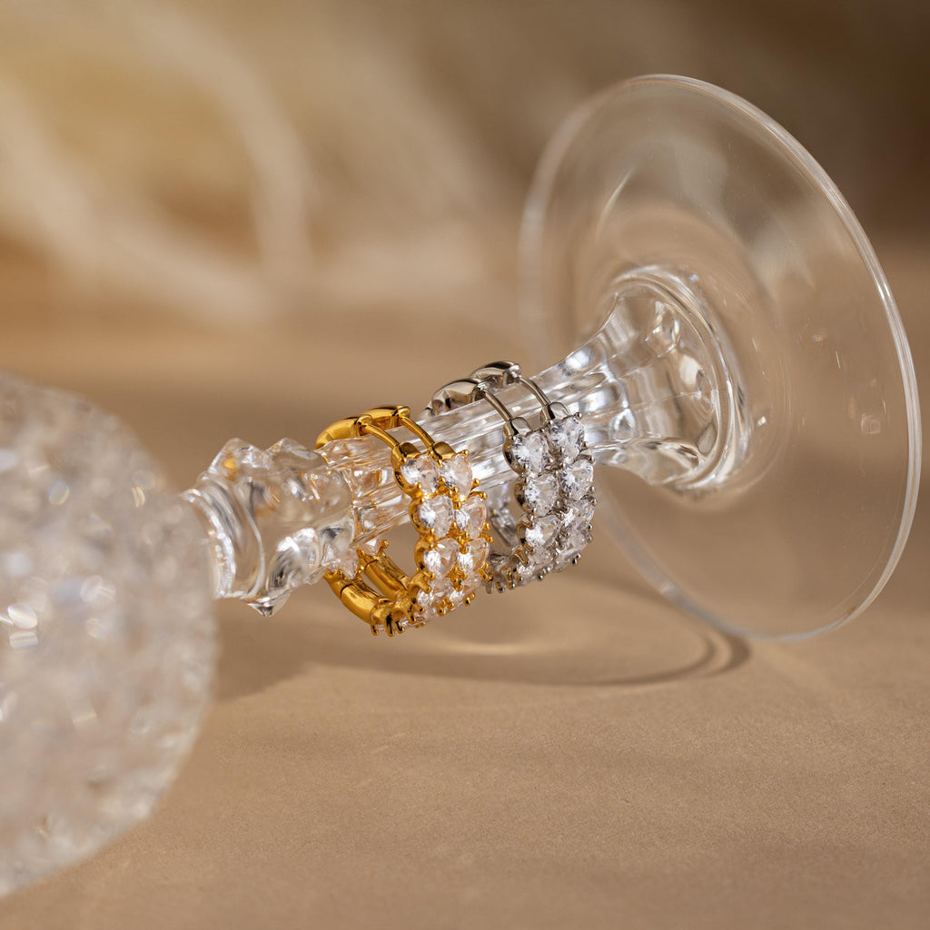 The Inside Out Heart Diamond Hoops, inspired by heart-shaped diamond earrings, feature crystals in gold and silver finishes and are displayed on the stem of an upside-down wine glass.