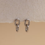 The Link Drop Studs are elegant earrings with a refined double-link design, shown on a textured beige surface against a neutral backdrop.