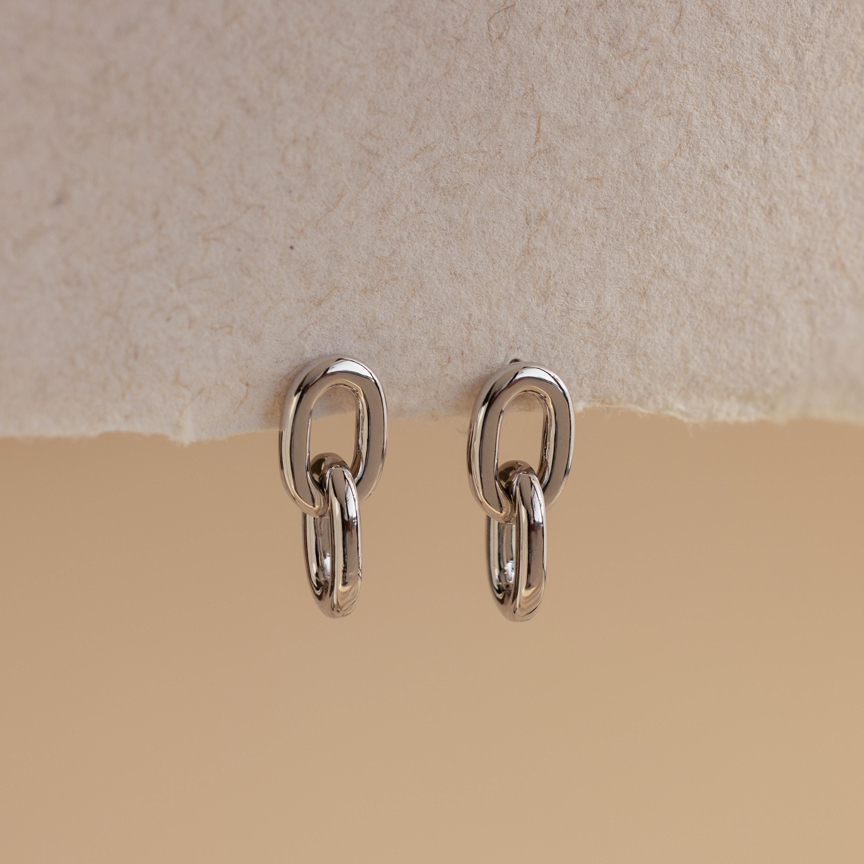 The Link Drop Studs are elegant earrings with a refined double-link design, shown on a textured beige surface against a neutral backdrop.