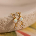 Opal & Diamond Drop Studs, featuring gold with opal and diamond accents, are elegantly displayed on a textured cream-colored surface.