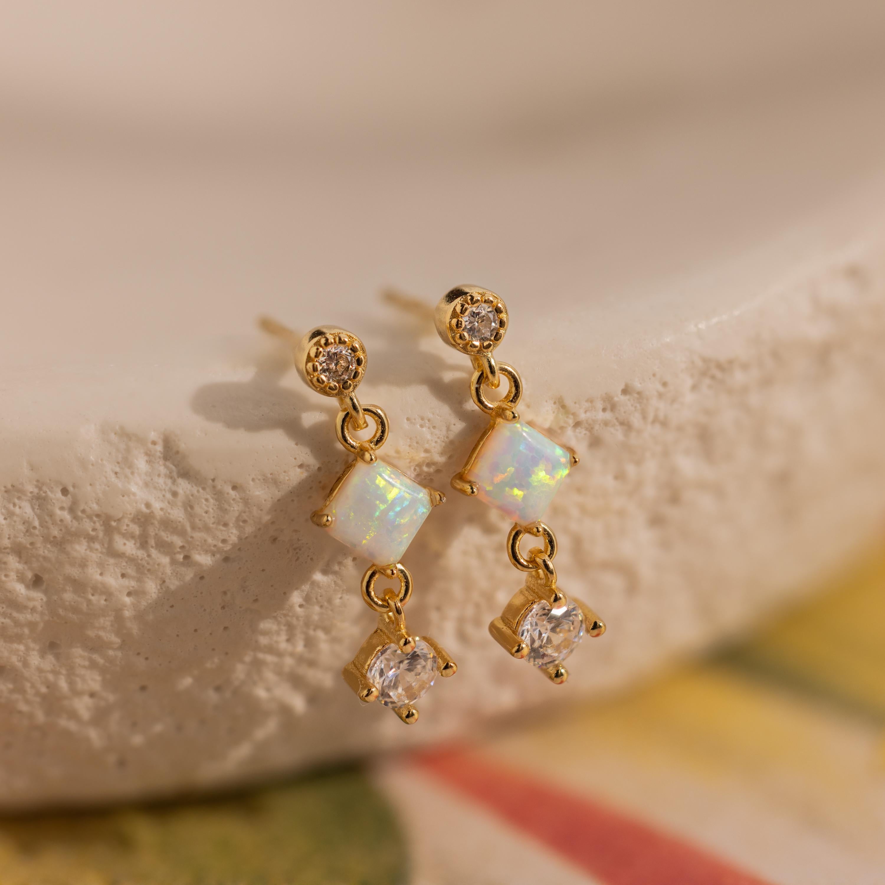 Opal & Diamond Drop Studs, featuring gold with opal and diamond accents, are elegantly displayed on a textured cream-colored surface.