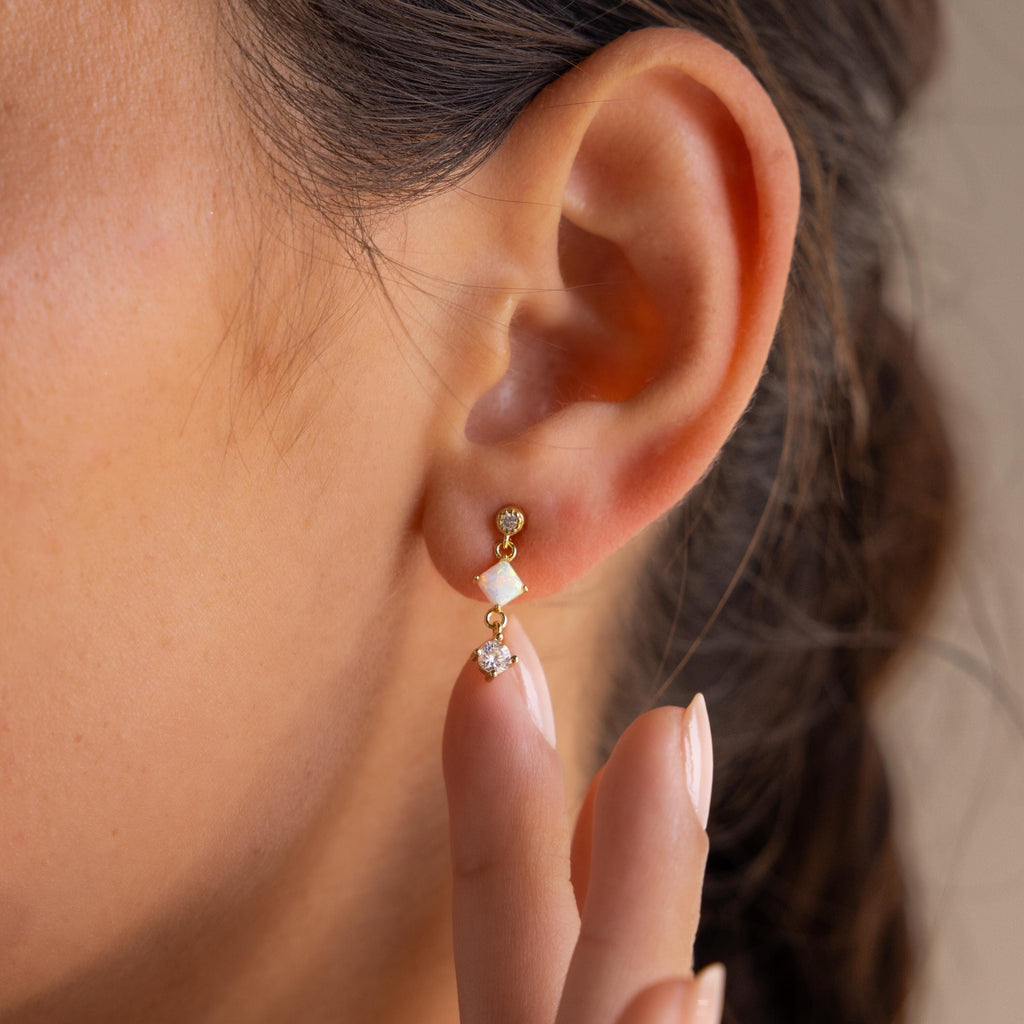 A close-up of a woman's ear wearing Opal & Diamond Drop Studs in gold and white, with her finger gently touching her earlobe.