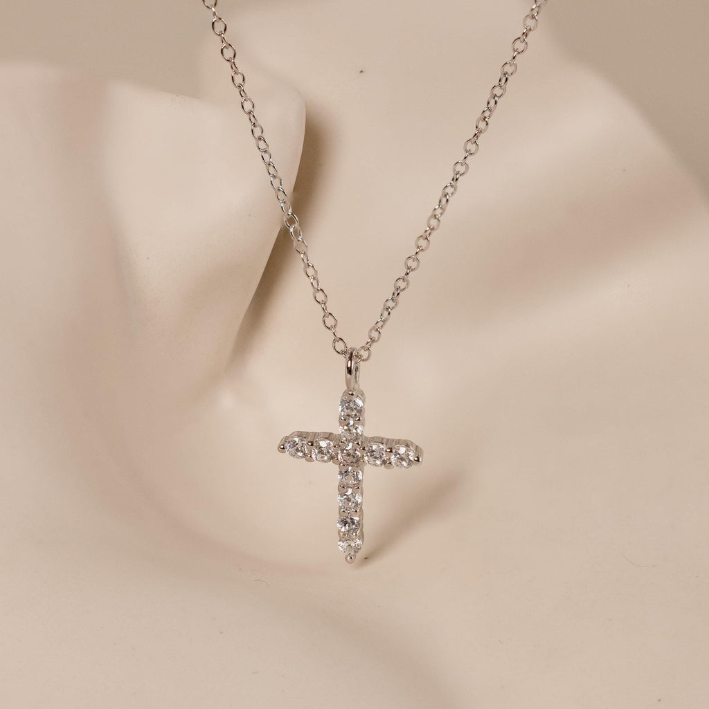 The Pave Cross Necklace features a silver chain and rhinestone cross pendant, elegantly displayed on a neutral mannequin bust.